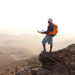 A person wearing a sun hat and orange backpack holding a map. They're standing on a mountain looking down at the scenic view below.