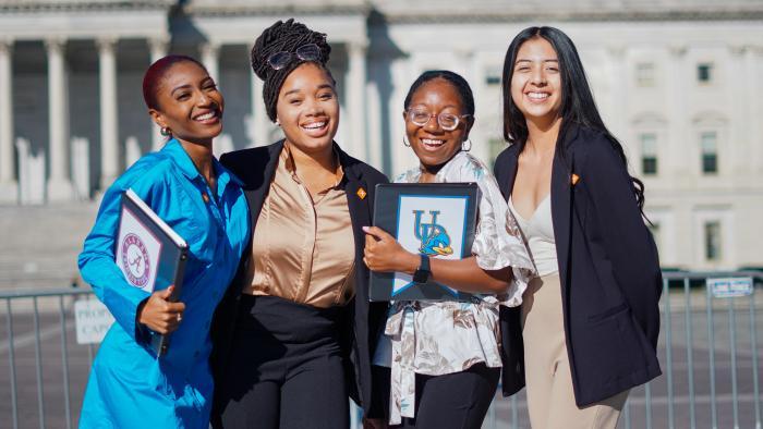 Four smiling young women stand together outside a government building, holding folders and wearing business attire, as they pose confidently in bright sunlight.