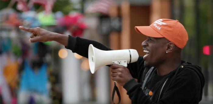 A man wearing an orange cap speaks into a megaphone and points into the distance while standing outdoors in a lively urban setting.