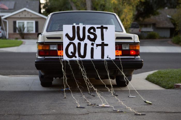 Rear of car displaying “JUST QUIT” sign with strings holding vapes trailing onto suburban street.