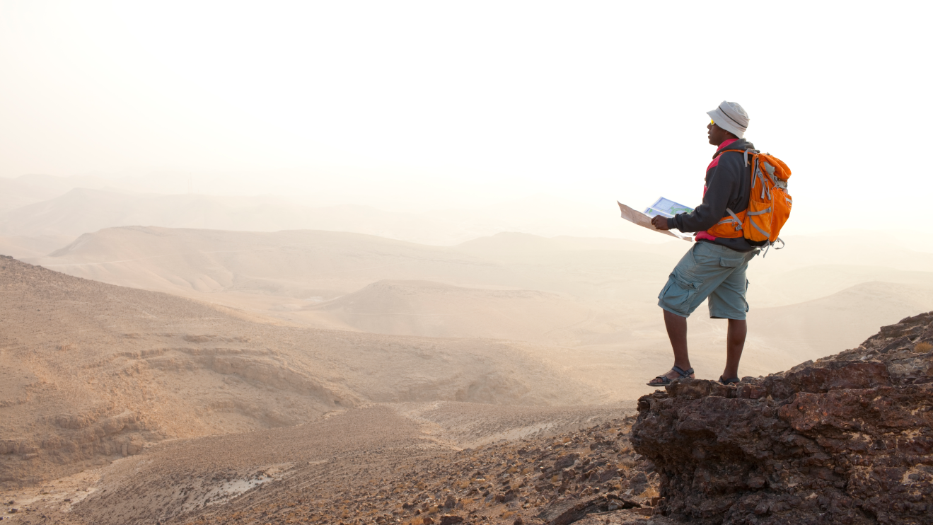 A person wearing a sun hat and orange backpack holding a map. They're standing on a mountain looking down at the scenic view below.