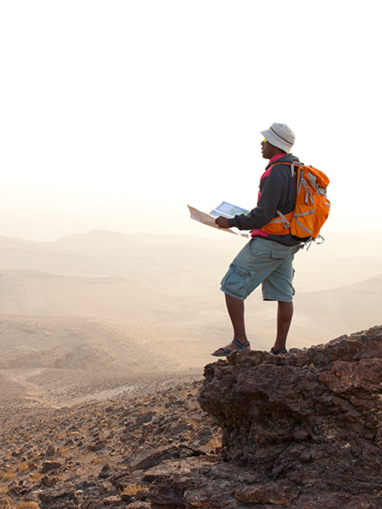 A person wearing a sun hat and orange backpack holding a map. They're standing on a mountain looking down at the scenic view below.