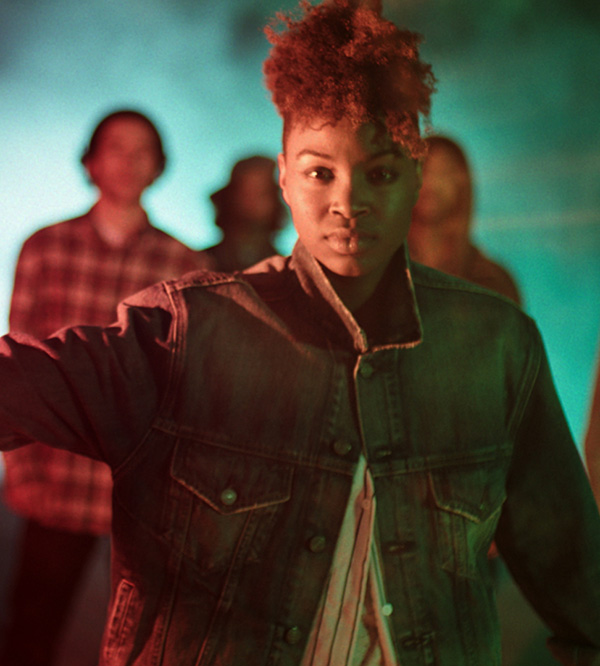 	A young person with short reddish hair wears a dark green jacket over a red shirt, standing in a group under moody teal and orange lighting.