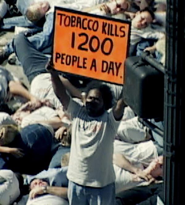 A person stands holding an orange protest sign reading "TOBACCO KILLS 1200 PEOPLE A DAY" above a crowd of people lying on the ground in a die-in demonstration.