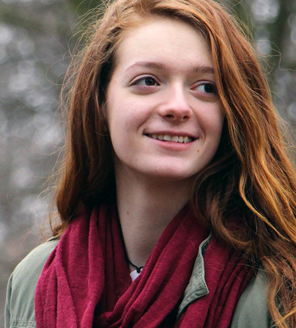 A smiling young woman with long red hair wears a dark red scarf, photographed outdoors against a blurred wintry background of bare trees.