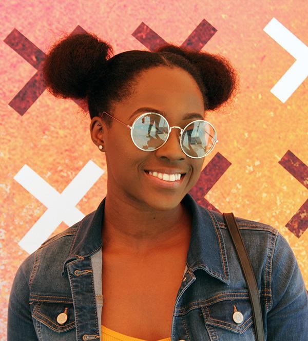 A smiling young woman with space buns and round mirrored sunglasses wears a denim jacket, standing in front of an orange and pink wall patterned with white X marks.