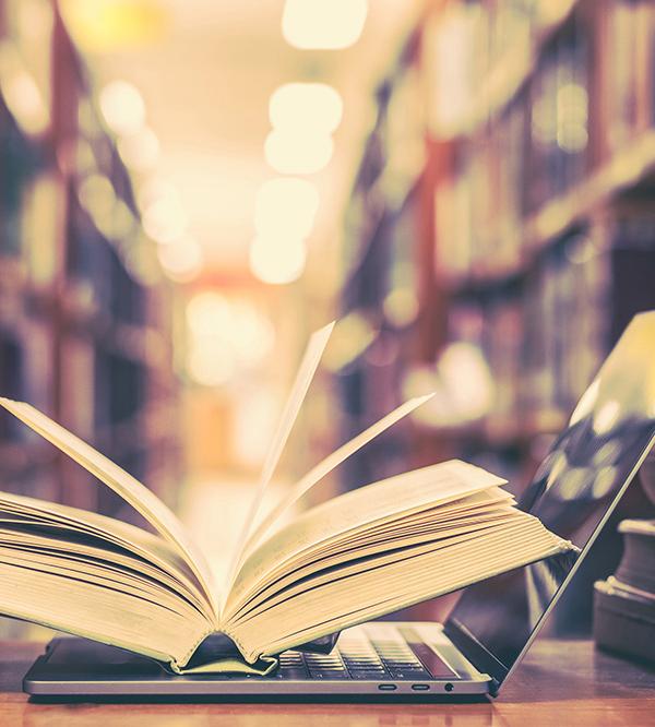An open book resting on an open laptop in a library aisle, with rows of bookshelves blurred in the warm, softly lit background.