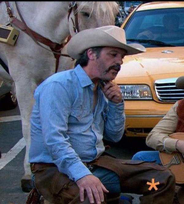 A man in a tan cowboy hat and blue denim shirt kneels on a city street beside a white horse and a yellow taxi